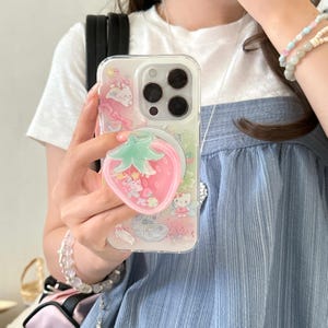 May include: Clear phone case with a pink strawberry-shaped phone grip. The case features a colorful design with cartoon characters and pink accents. The phone case is designed to fit a smartphone.