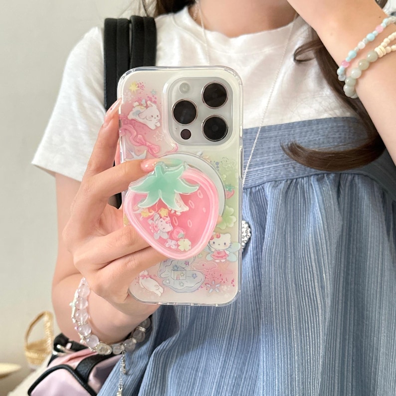 May include: Clear phone case with a pink strawberry-shaped phone grip. The case features a colorful design with cartoon characters and pink accents. The phone case is designed to fit a smartphone.