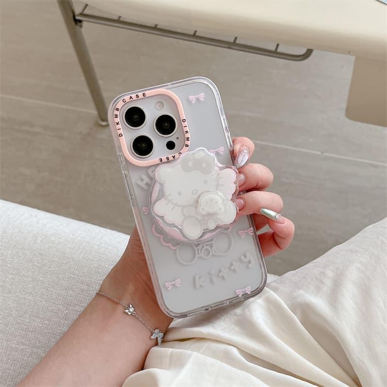 K&ouml;nnte beinhalten: Transparente Handyh&uuml;lle mit rosa Rand und wei&szlig;em Hello Kitty-Design. Die H&uuml;lle hat einen Pop-up-Griff mit einem Hello Kitty- und Teddyb&auml;r-Design. Auf der H&uuml;lle ist der Text "DIKMB CASE" und "Happy Kitty" aufgedruckt.