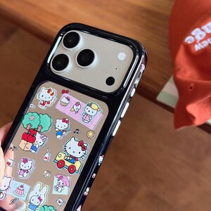 May include: Black phone case with a clear panel featuring colorful Hello Kitty stickers. The stickers include images of Hello Kitty, cupcakes, trees, and other cute designs. The case has a black border and a camera cutout.