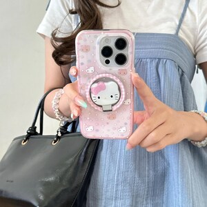 May include: A pink phone case with a Hello Kitty design. The case has a clear back with a pattern of Hello Kitty faces and stars. A matching pop-socket with a Hello Kitty face is attached. The phone case is held by a person.