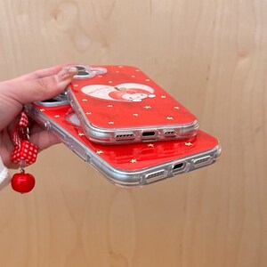 Puede incluir: Dos fundas rojas para tel&eacute;fono con borde transparente apiladas. La funda superior presenta un dise&ntilde;o en forma de coraz&oacute;n con un personaje de manzana de dibujos animados y detalles de estrellas. Se adjunta una correa de mu&ntilde;eca a cuadros rojos y blancos con un dije de cereza.