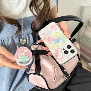 May include: A clear phone case with a colorful cartoon design is held near a pink purse with black trim. A small, pink strawberry-shaped compact mirror is also visible. A silver heart pendant hangs from a necklace.