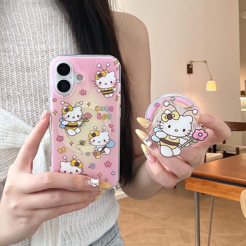 May include: Pink phone case with a cartoon design featuring Hello Kitty dressed as a bee, with the text "Cute Bee". A matching phone grip is also visible, with a similar design. The case is on a phone.