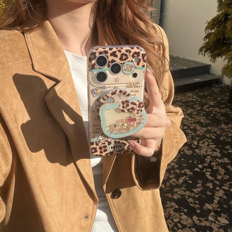 May include: Clear phone case with a leopard print design and a pop-up grip. The case features a cartoon character and the word "KITTY". The grip has a light blue border and contains small, colorful objects.