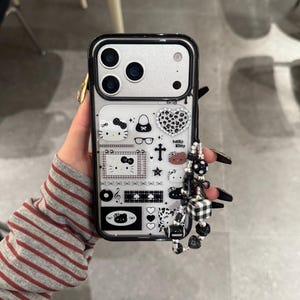 Op de afbeelding: Zwarte en zilveren telefoonhoesje met een Hello Kitty-ontwerp en diverse iconen. Het hoesje heeft een kralenbedel met zwart-witte accenten. Telefoonaccessoire.