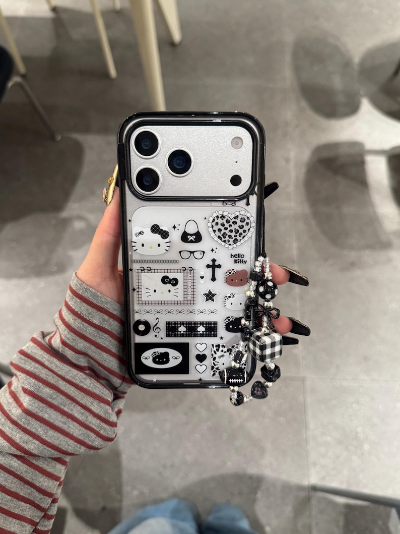 Op de afbeelding: Zwarte en zilveren telefoonhoesje met een Hello Kitty-ontwerp en diverse iconen. Het hoesje heeft een kralenbedel met zwart-witte accenten. Telefoonaccessoire.