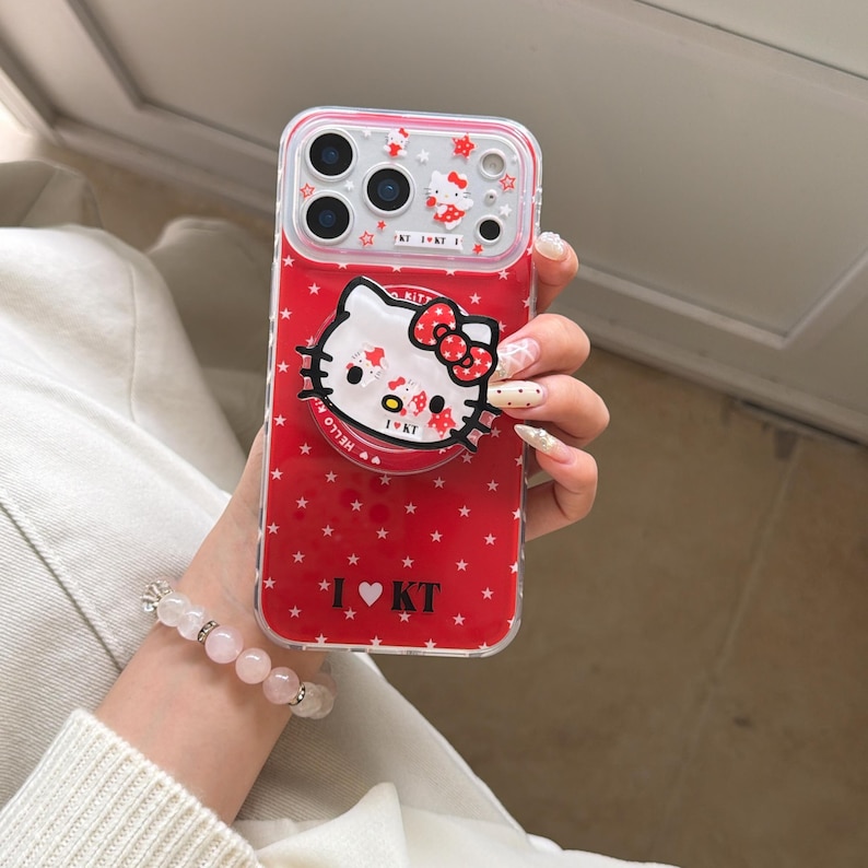 May include: Red phone case with a Hello Kitty design. The case has a pop-up grip featuring Hello Kitty's face and a red bow. The case is decorated with white stars and the text "I ❤️ KT".