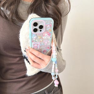 Puede incluir: Una funda transparente para teléfono con un borde azul claro y rosa con personajes de dibujos animados. La funda tiene una correa decorativa de cuentas con cuentas de colores pastel y un dije rosa.