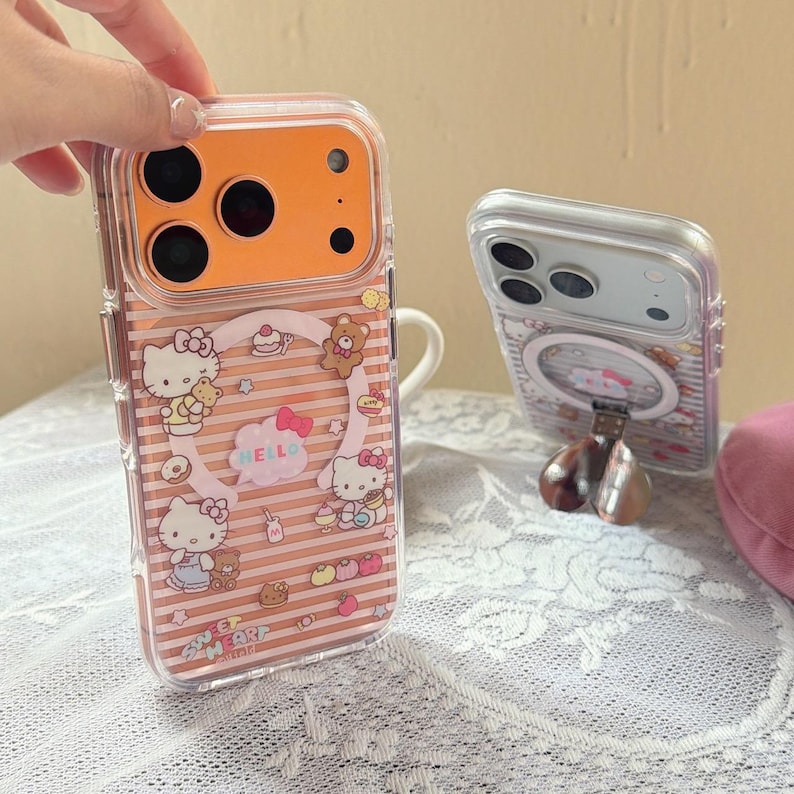 May include: Clear phone case with a pink and brown striped design featuring Hello Kitty characters, bears, and the text "HELLO" and "SWEET HEART". The case has an orange camera section and is held by a hand.
