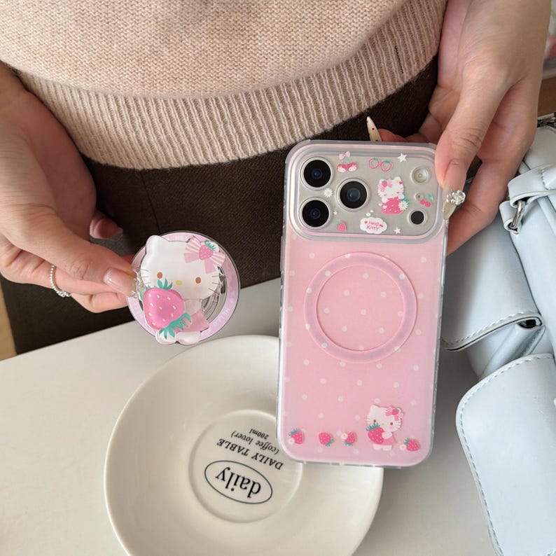 May include: A pink phone case with a white polka dot pattern and a Hello Kitty design. The case has a clear border and a circular magnetic attachment area. A matching phone grip with a Hello Kitty and strawberry design is also visible.