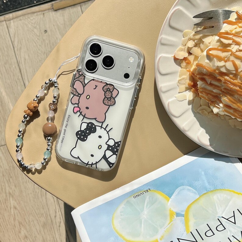 May include: Clear phone case with a cartoon design featuring a pink and white cat character. The case has a wrist strap with beads. The text "enjoy the good times" is printed on the case. A slice of cake with almonds and caramel is on a white plate.