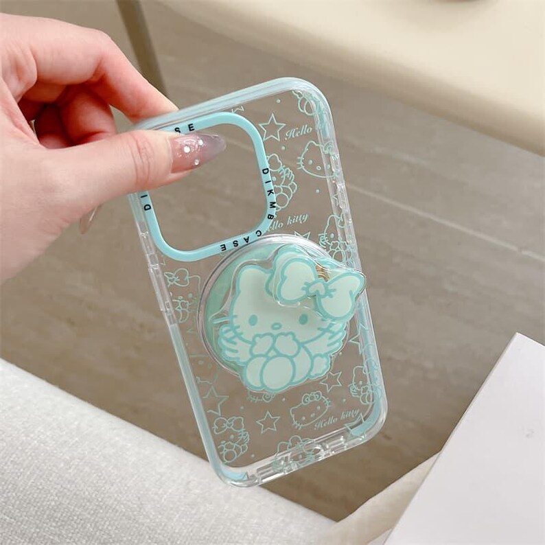 K&ouml;nnte beinhalten: Transparente Handyh&uuml;lle mit hellblauem Rahmen und passendem Pop-Socket mit Hello Kitty-Design. Die H&uuml;lle hat ein sich wiederholendes Muster aus Hello Kitty-Illustrationen und Sternen.