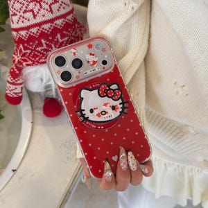 May include: Red phone case featuring a Hello Kitty design with a red bow and star pattern. The case has a clear border and includes the text "Hello Kitty" and "I ♡ KT".