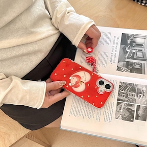 Puede incluir: Funda de tel&eacute;fono roja con un dise&ntilde;o de Hello Kitty, con un gr&aacute;fico en forma de coraz&oacute;n y estrellas doradas. La funda se sostiene sobre un libro abierto. Un peque&ntilde;o dije rojo con una cara de Hello Kitty y dados est&aacute; adjunto al tel&eacute;fono.