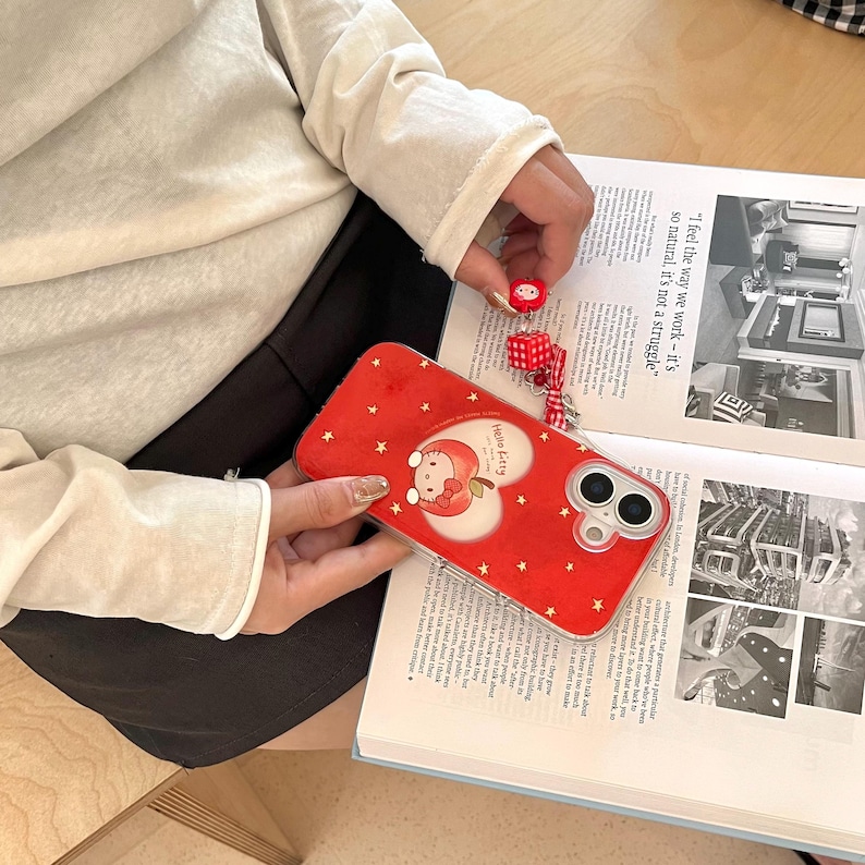 Puede incluir: Funda de tel&eacute;fono roja con un dise&ntilde;o de Hello Kitty, con un gr&aacute;fico en forma de coraz&oacute;n y estrellas doradas. La funda se sostiene sobre un libro abierto. Un peque&ntilde;o dije rojo con una cara de Hello Kitty y dados est&aacute; adjunto al tel&eacute;fono.