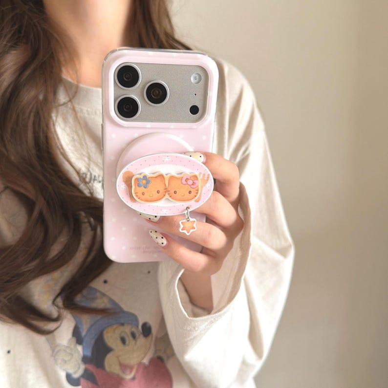 May include: A pink phone case with a pop-up grip featuring a cartoon design of two orange cats with bows. The case has a light pink polka dot pattern and a star-shaped charm hanging from the grip.