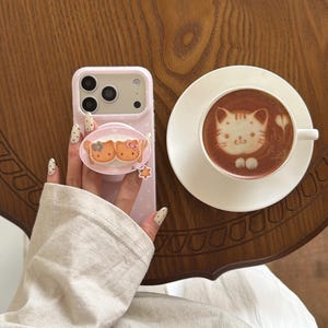 May include: A pink phone case with a pop-up grip featuring two cartoon cats. A cup of coffee with cat art is on a white saucer. The items are on a wooden table. The phone case has a polka dot design.