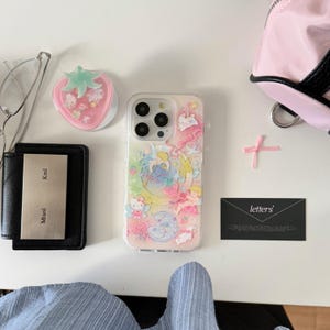 May include: A clear phone case with a colorful illustration of cartoon characters, a pink strawberry phone grip, a black wallet with a silver nameplate, and a black business card with the word "letters".