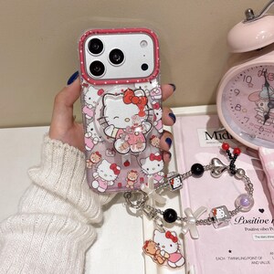 May include: A clear phone case with a pink border and a pop-up grip featuring Hello Kitty. The case is decorated with Hello Kitty and teddy bear graphics. A charm bracelet with Hello Kitty charms is attached.