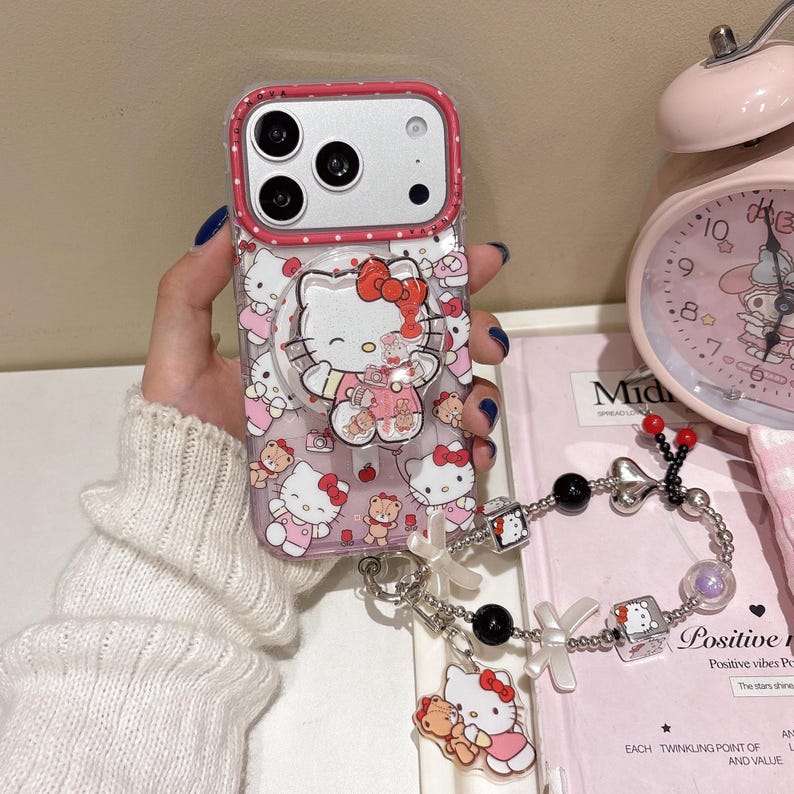 May include: A clear phone case with a pink border and a pop-up grip featuring Hello Kitty. The case is decorated with Hello Kitty and teddy bear graphics. A charm bracelet with Hello Kitty charms is attached.