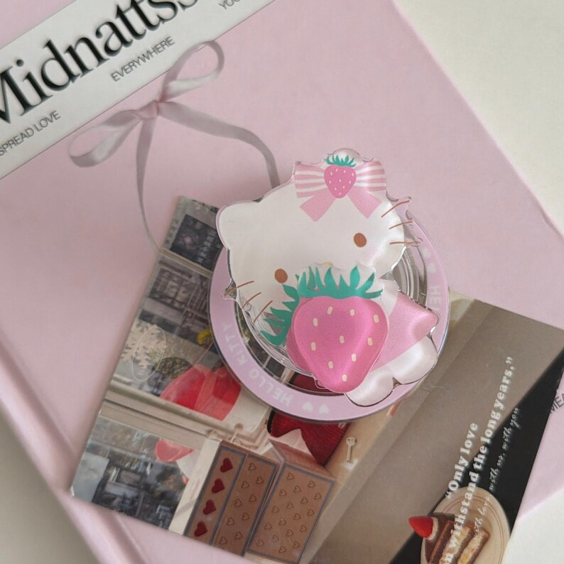 May include: A Hello Kitty pin featuring a strawberry design, set against a pink background with the text "Midnatts" and "Everywhere". The pin has a white face, pink bow, and a large pink strawberry. The pin is on top of a pink book.