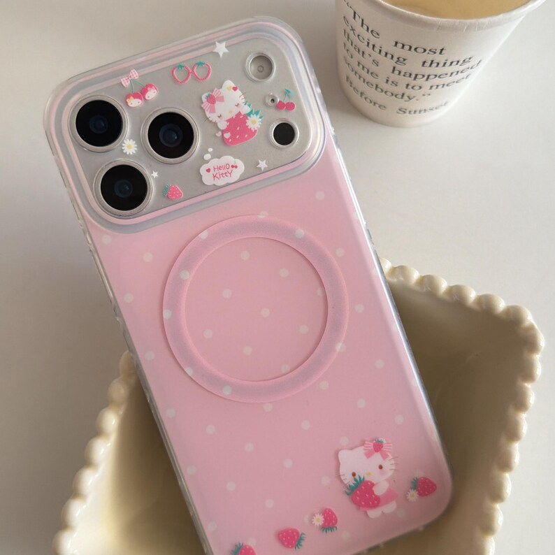May include: Pink phone case with a Hello Kitty design. The case features a pink polka dot pattern, a clear camera lens protector, and decorative strawberry and cherry accents. A Hello Kitty graphic is visible on the lower right.