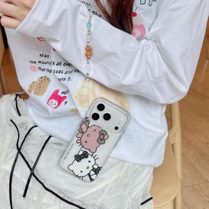 May include: Clear phone case with a Hello Kitty design, featuring two characters in pink and black. The case has a lanyard with colorful beads. The person is wearing a white long-sleeve shirt with text and illustrations.