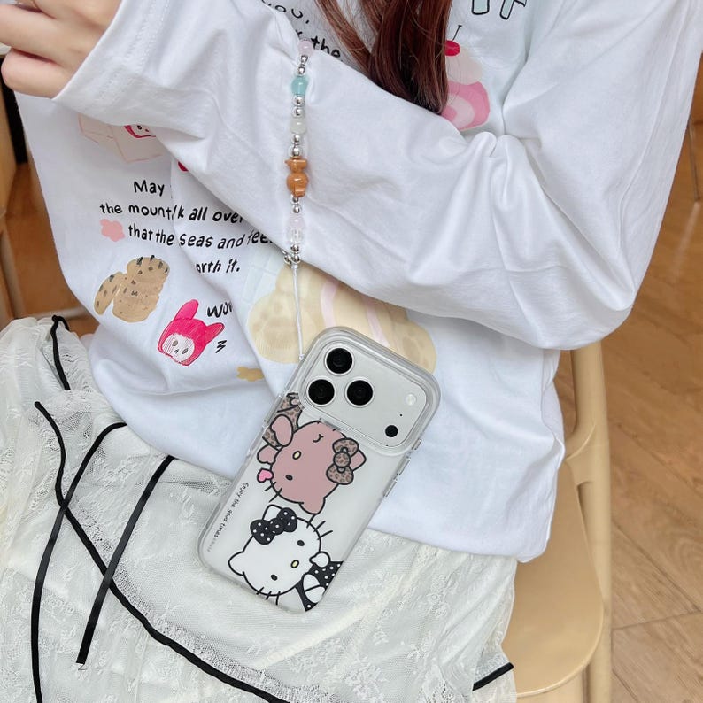 May include: Clear phone case with a Hello Kitty design, featuring two characters in pink and black. The case has a lanyard with colorful beads. The person is wearing a white long-sleeve shirt with text and illustrations.