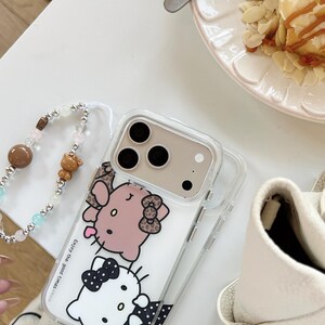 May include: Clear phone case featuring a cartoon design of a pink cat with a bow and a white cat with a black bow. The case has a beaded wrist strap with a brown bead and a small bear charm. The text "Enjoy the good times" is printed on the case.