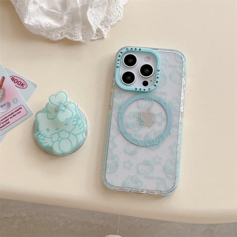 K&ouml;nnte beinhalten: Eine transparente Handyh&uuml;lle mit hellblauem Sto&szlig;f&auml;nger und passendem Pop-Socket. Die H&uuml;lle hat ein sich wiederholendes Muster von Zeichentrickfiguren und einen hellblauen Ring f&uuml;r ein magnetisches Ladeger&auml;t. Der Pop-Socket hat ein Zeichentrickfiguren-Design.