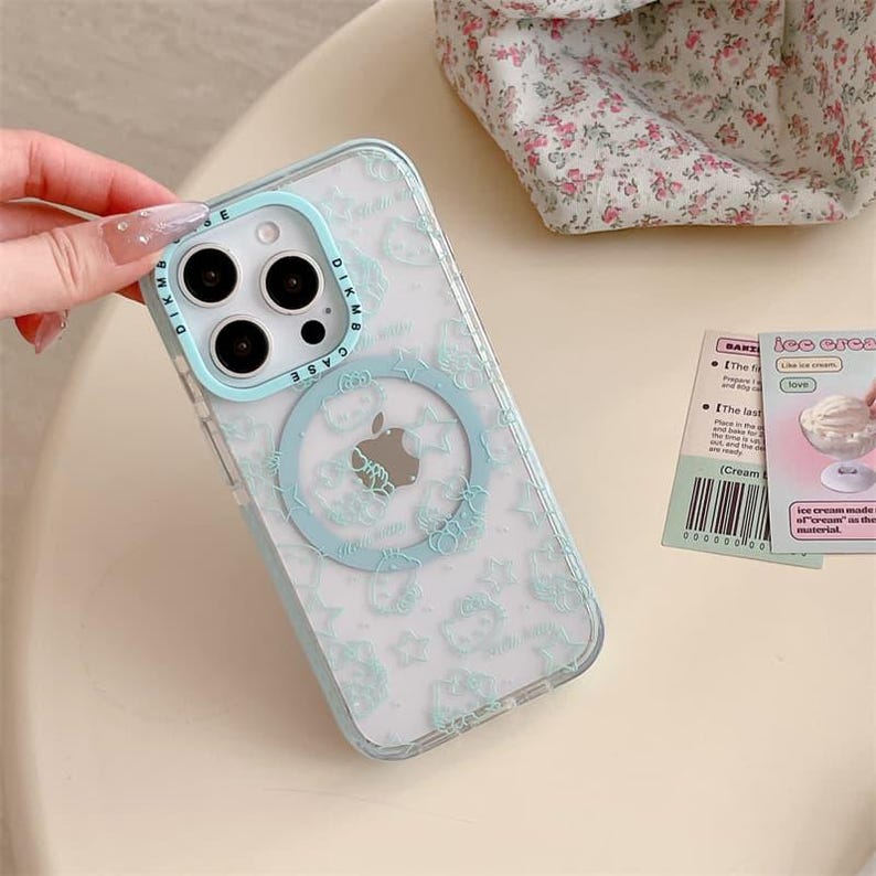 K&ouml;nnte beinhalten: Transparente iPhone-H&uuml;lle mit hellblauem Rand und passendem kreisf&ouml;rmigen Design. Die H&uuml;lle zeigt ein Muster aus Zeichentrickfiguren und Sternen. Die H&uuml;lle wird von einer Hand mit manik&uuml;rten N&auml;geln gehalten. Die H&uuml;lle hat den Text "DIKMB CASE".