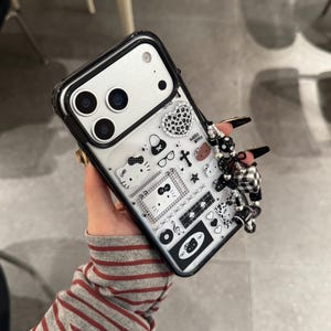 Op de afbeelding: Zwarte en doorzichtige telefoonhoes met een Hello Kitty-ontwerp. De hoes heeft een verscheidenheid aan kleine, zwart-witte afbeeldingen, waaronder harten, strikken en een kruis. Een zwart-wit geruite bedel is aan de zijkant bevestigd.