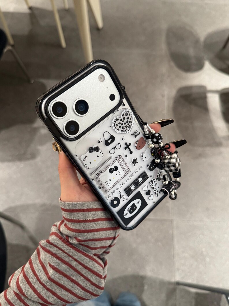 Op de afbeelding: Zwarte en doorzichtige telefoonhoes met een Hello Kitty-ontwerp. De hoes heeft een verscheidenheid aan kleine, zwart-witte afbeeldingen, waaronder harten, strikken en een kruis. Een zwart-wit geruite bedel is aan de zijkant bevestigd.