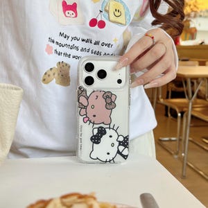 May include: Clear phone case featuring a pink and white cartoon character design. The case has a transparent background and includes the text "Enjoy the good times". The phone case is held by a person.