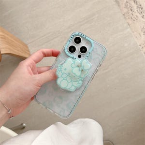 K&ouml;nnte beinhalten: Transparente Handyh&uuml;lle mit hellblauem Rand und hellblauem Hello Kitty-Design. Die H&uuml;lle hat ein Blumenmuster und den Text "DIKM CASE". Die Handyh&uuml;lle wird von einer Person gehalten.