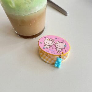 Puede incluir: Un soporte para tel&eacute;fono rosa y amarillo con dos personajes de Hello Kitty, estrellas y el texto "Have a nice day". Un peque&ntilde;o dije de osito de goma azul cuelga del soporte. Un vaso de matcha latte helado est&aacute; en el fondo.