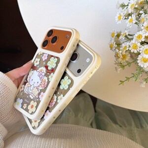 May include: Two phone cases with a floral and Hello Kitty design. The cases have a cream-colored border with a brown camera section. One case features Hello Kitty surrounded by flowers, while the other has a floral pattern.