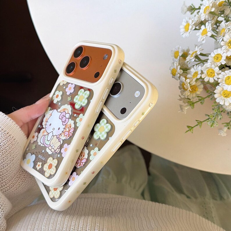 May include: Two phone cases with a floral and Hello Kitty design. The cases have a cream-colored border with a brown camera section. One case features Hello Kitty surrounded by flowers, while the other has a floral pattern.