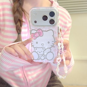 May include: Clear phone case with a Hello Kitty design, pink bow, and small teddy bear. The case has a silver glitter border and a pink beaded charm with heart and bow details.