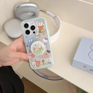 May include: A clear phone case with a mint green border, decorated with cartoon characters and a pop socket with floating glitter. The case is held near silver headphones and a light blue box.