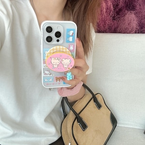 Puede incluir: Funda transparente para tel&eacute;fono con un dise&ntilde;o colorido con personajes de dibujos animados. La funda tiene un agarre emergente con un patr&oacute;n rosa y amarillo y un dije de osito de goma azul. La funda del tel&eacute;fono se sostiene frente a una camisa blanca y un bolso de mano marr&oacute;n.