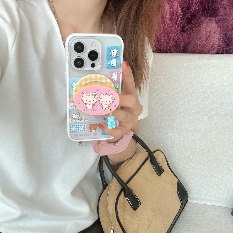 Puede incluir: Funda transparente para tel&eacute;fono con un dise&ntilde;o colorido con personajes de dibujos animados. La funda tiene un agarre emergente con un patr&oacute;n rosa y amarillo y un dije de osito de goma azul. La funda del tel&eacute;fono se sostiene frente a una camisa blanca y un bolso de mano marr&oacute;n.