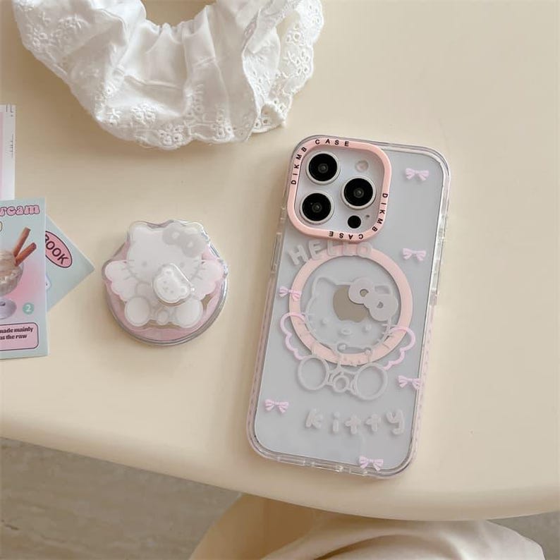 K&ouml;nnte beinhalten: Eine transparente Handyh&uuml;lle mit rosa Rand und Hello Kitty-Design. Die H&uuml;lle hat ein rosa Schleifenmuster und das Wort "Kitty" unten. Ein passender Handyhalter und eine wei&szlig;e Spitzenscrunchie sind ebenfalls sichtbar.