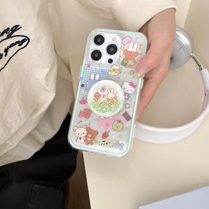 May include: A clear phone case with a light blue border, decorated with cartoon characters and a pop-up grip. The case displays colourful illustrations of bunnies, bears, and other cute designs. The pop-up grip has a glittery, green background.