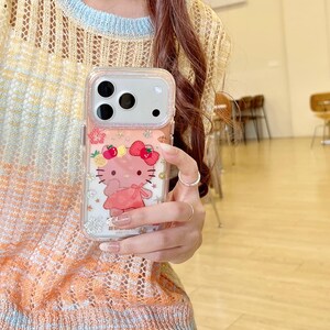 May include: Clear phone case featuring a Hello Kitty design. The case has a pink background with a Hello Kitty graphic wearing a pink dress and a bow. The case is held by a person wearing a light orange and yellow sweater.