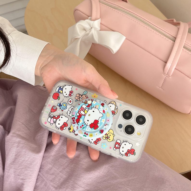 May include: Clear phone case featuring a colorful Hello Kitty design. The case has a pop-up grip with a Hello Kitty graphic. The case is held in a hand, with a pink bag and white bow in the background.
