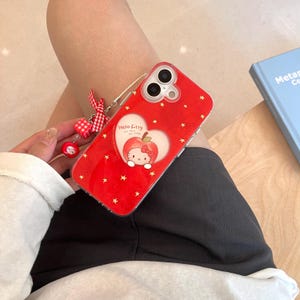 Puede incluir: Funda de tel&eacute;fono roja con un dise&ntilde;o de manzana de Hello Kitty y detalles de estrellas doradas. La funda tiene una correa transparente y un charm a juego. El texto "Hello Kitty" es visible.