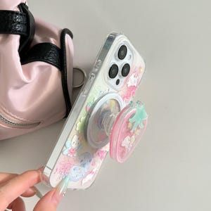 May include: Clear phone case with a colorful design featuring cartoon characters and a pink strawberry-shaped phone grip. The case is attached to a phone with a triple-lens camera.