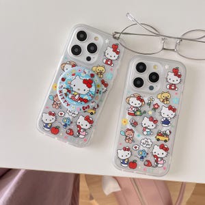 May include: Two clear phone cases with Hello Kitty designs. One case has a pop socket with a Hello Kitty graphic and the text "Kitty". The other case features various Hello Kitty illustrations with flowers and other characters.